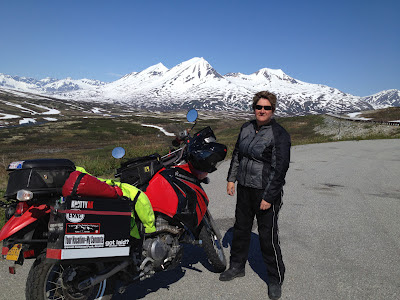 Judy in full gear standing beside her motorcycle with snow capped mountains in the distance