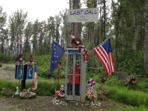 Image of phone booths in the middle of nowhere with lots of flags, flowers, and a sign reading, "Last Call 25 miles Haines, Alaska"